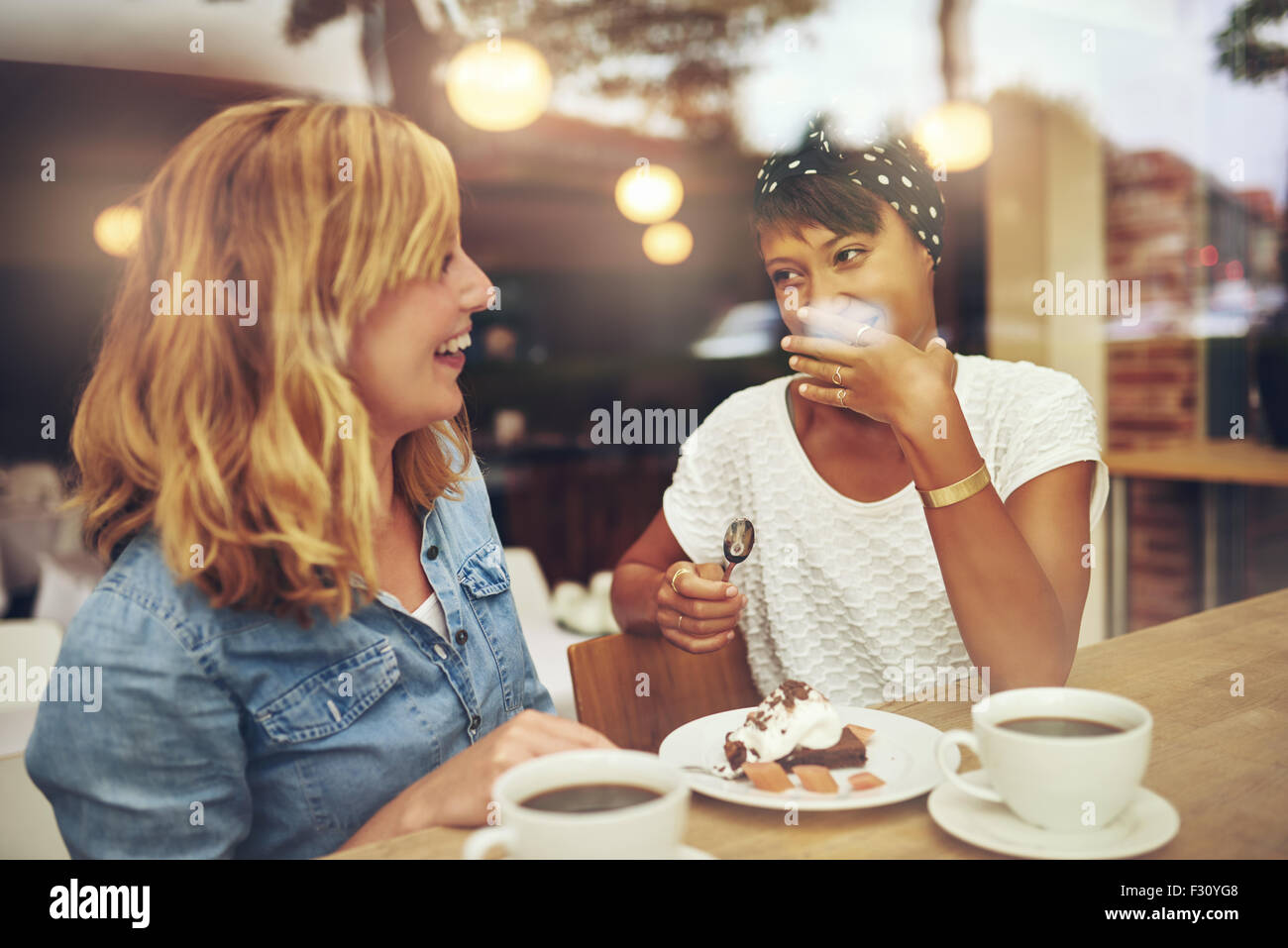 Two good female friends enjoying a cup of coffee together in a coffee house with one laughing as the other makes a gesture of em Stock Photo