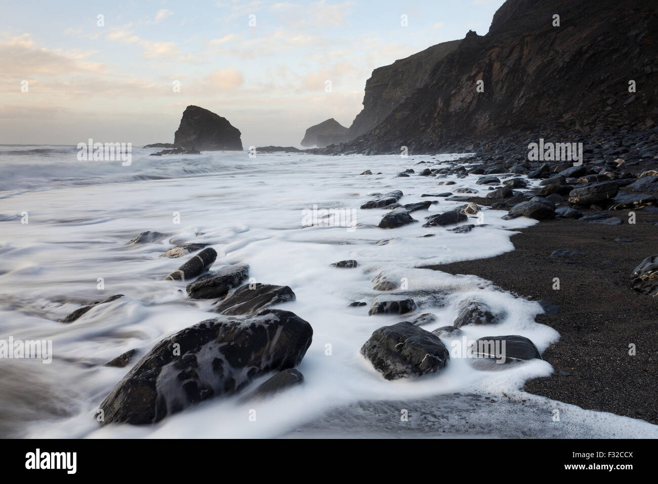 View of sea cliffs and beach with tide washing back over rocky black shingle beach on windy evening, The Strangles, North Cornwall, England, March Stock Photo