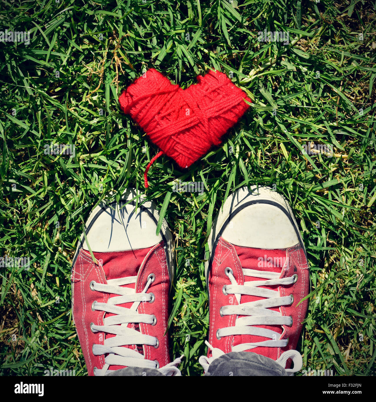 a heart-shaped coil of red rope and the feet of a man wearing red sneakers stepping on the grass, with a slight vignette added Stock Photo
