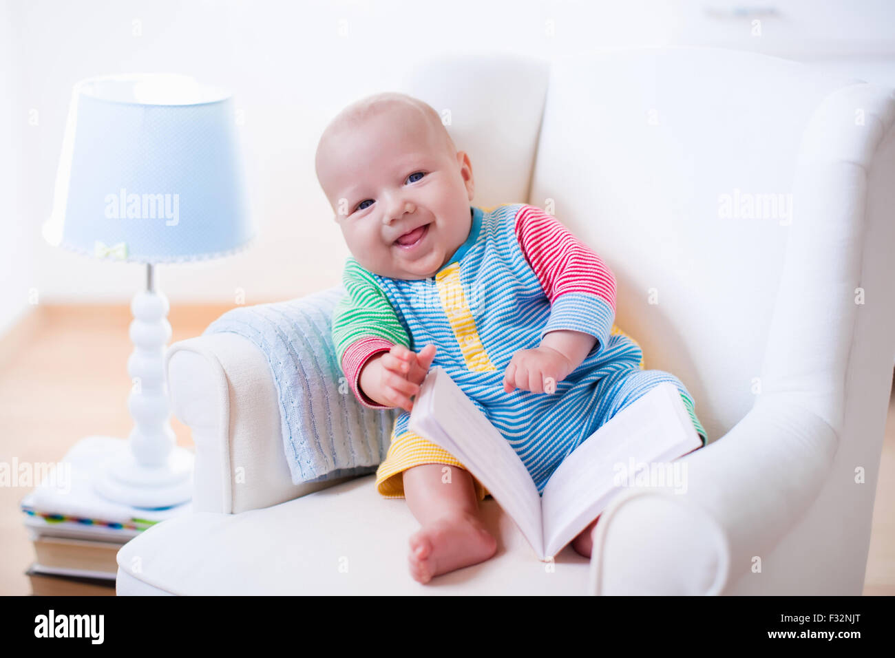 Cute funny baby boy reading a book sitting in a white chair at home