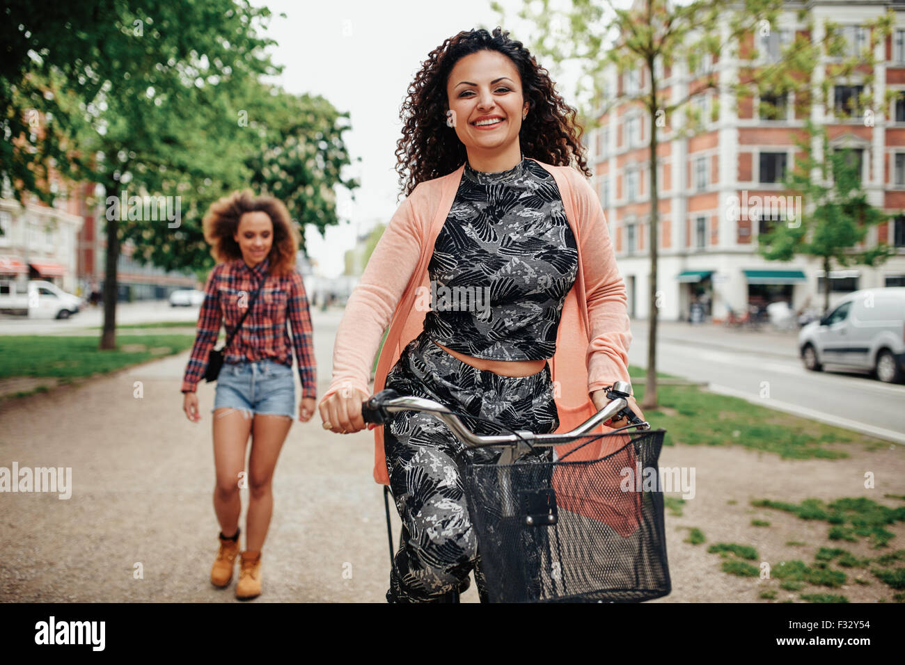 Portrait of happy young woman riding bicycle with another walking in background on city street. Stock Photo