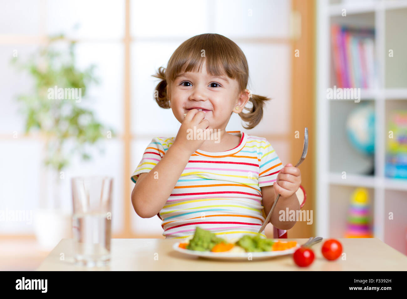 Happy child girl eats vegetables sitting at table in nursery Stock Photo