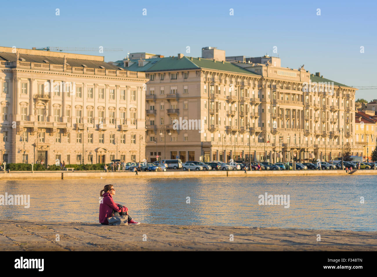 Trieste Italy harbor, view at sunset of a young woman sitting alone on the huge stone jetty (Molo Audace) in Trieste harbour, Italy Stock Photo