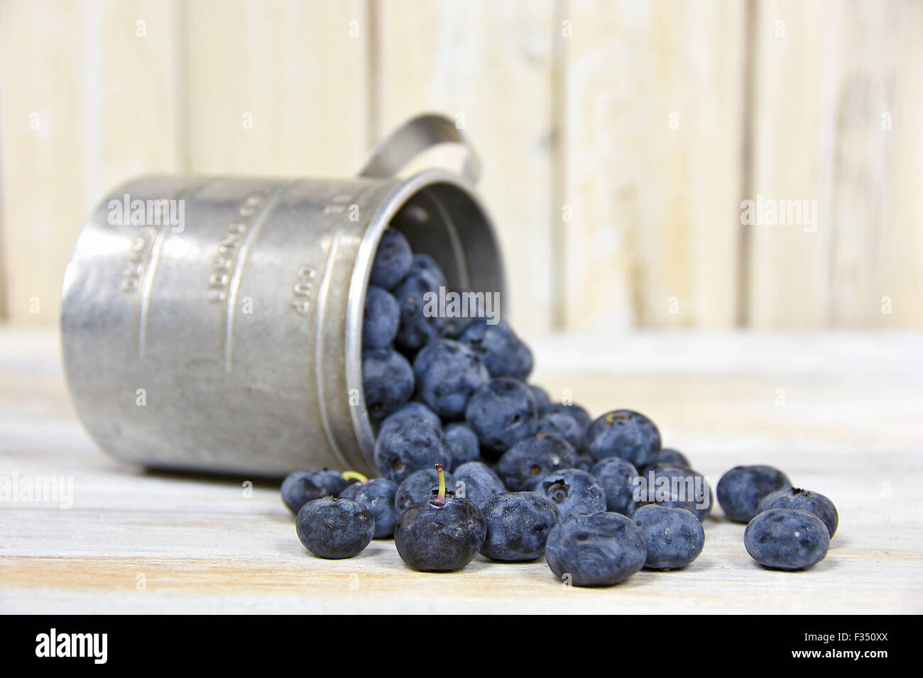 Ripe blueberries spilling out of retro tin measuring cup on whitewashed wood. Stock Photo