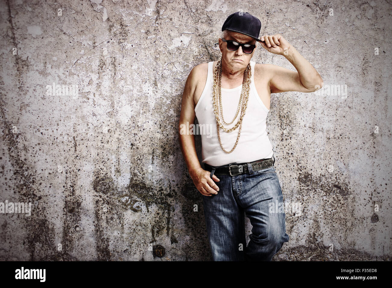 Senior gangster in hip hop outfit leaning against a rusty gray wall and looking at the camera Stock Photo
