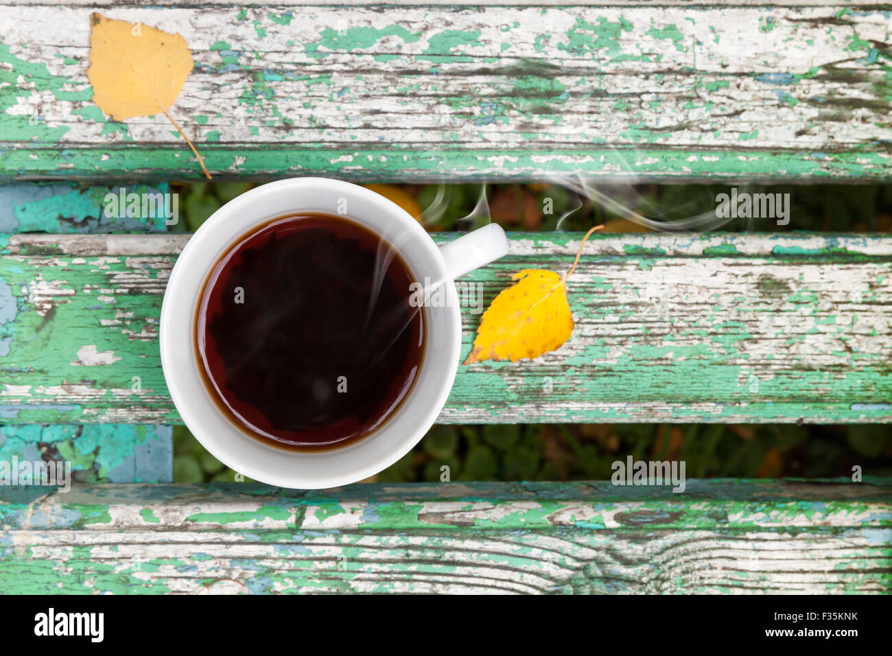White cup of black tea is on old green wooden bench in autumn park.  Selective focus with shallow DOF Stock Photo