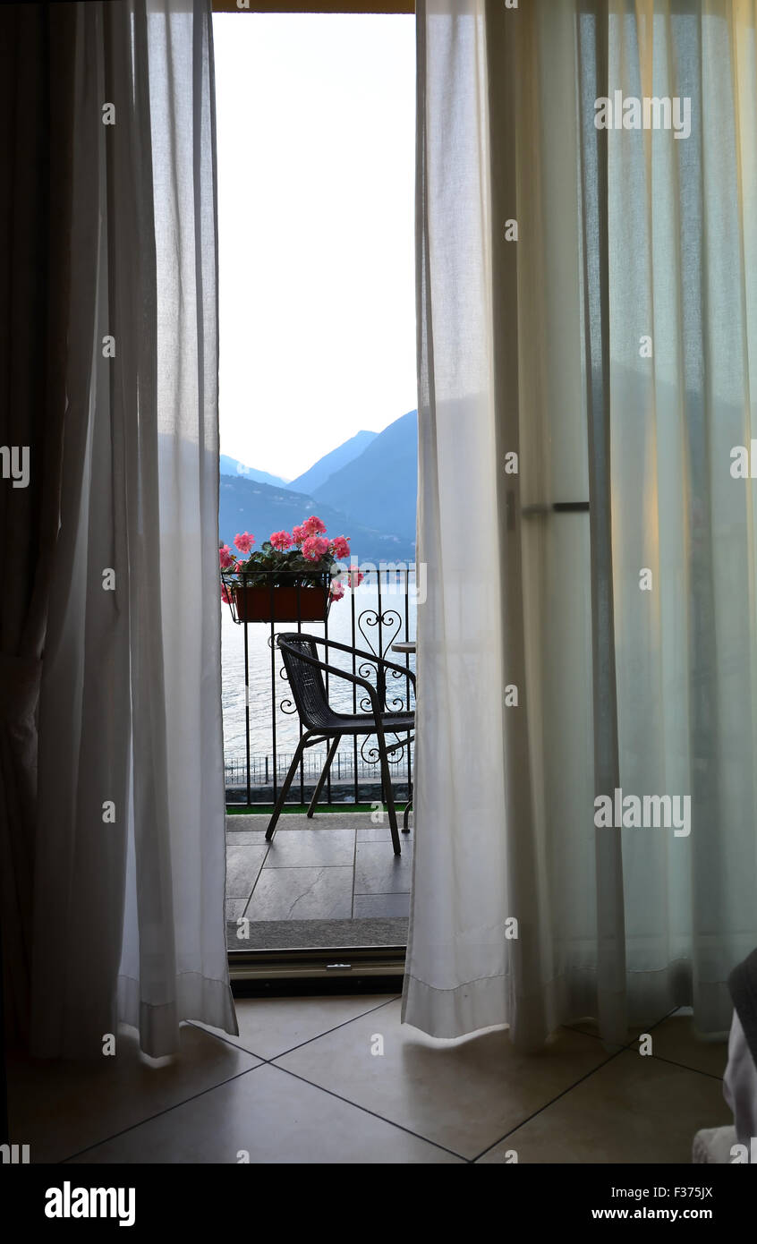 a gentle breeze wafts the curtains of a hotel room with balcony. through the open windows a romantic view of a lake in the mount Stock Photo
