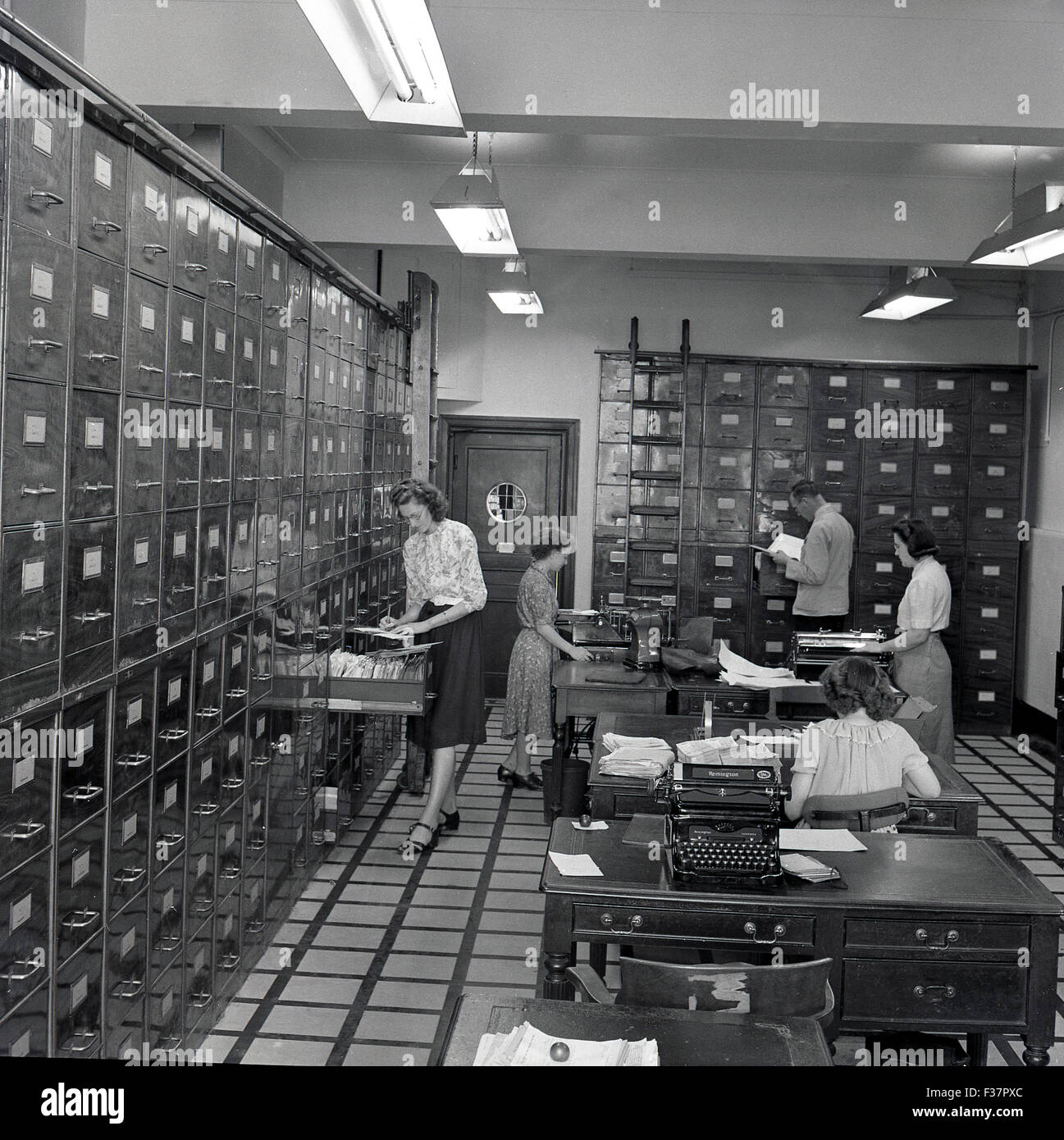 Historical, 1950s, female staff working in a large room filled with ceiling high shiny metal filing cabinets, several desks and typewriters. Stock Photo