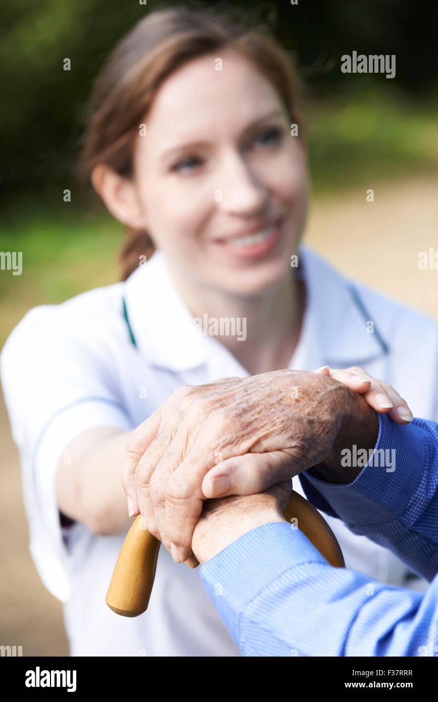 Senior Man's Hands Resting On Walking Stick With Care Worker In Background Stock Photo