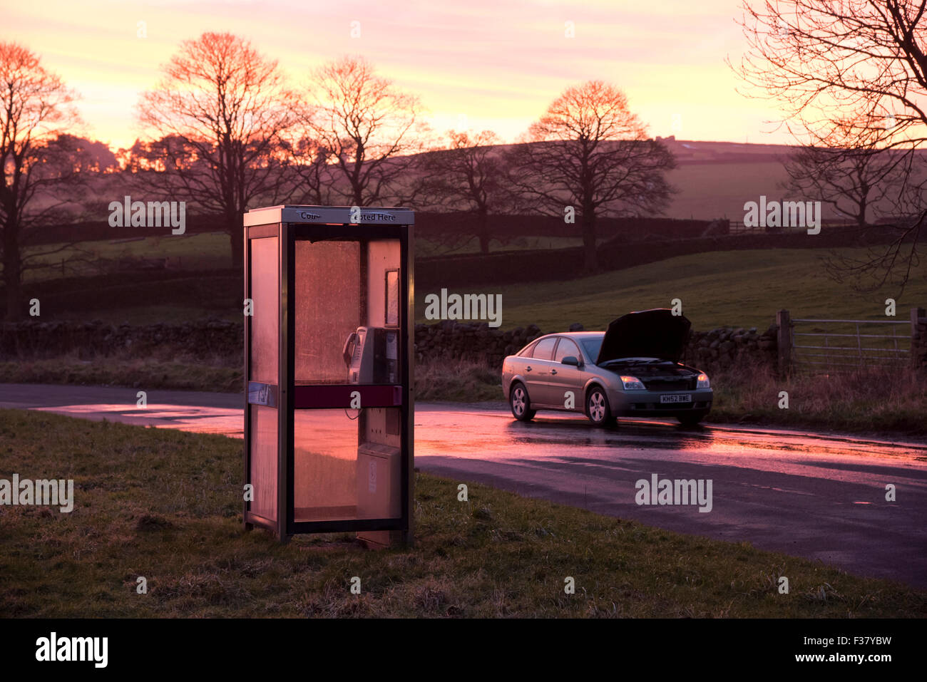 A car, bonnet raised after breaking down, parked at sunset in a deserted countryside lane (lay-by) alongside a public telephone box - England, UK. Stock Photo