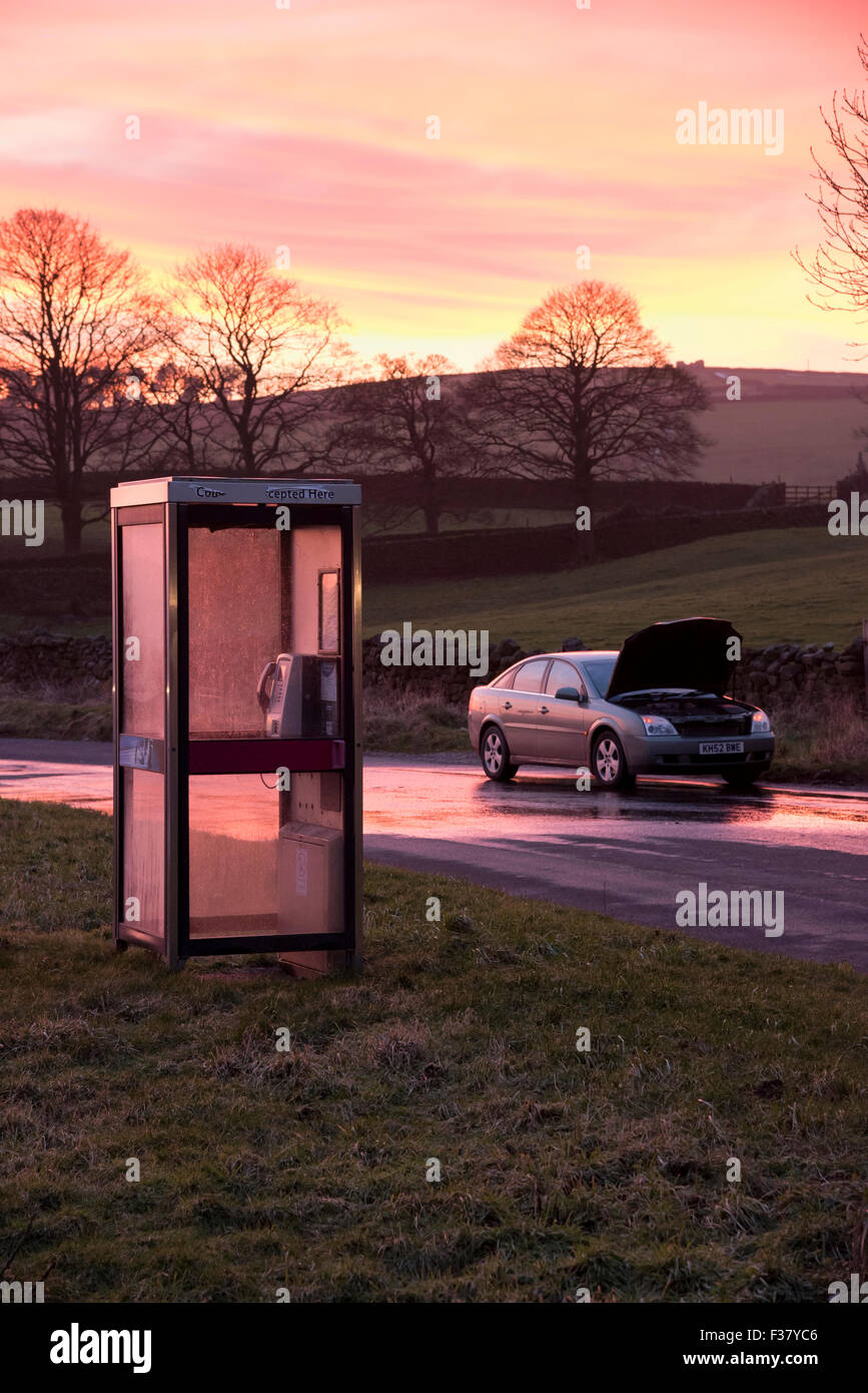 A car, bonnet raised after breaking down, parked at sunset in a deserted countryside lane (lay-by) alongside a public telephone box - England, UK. Stock Photo