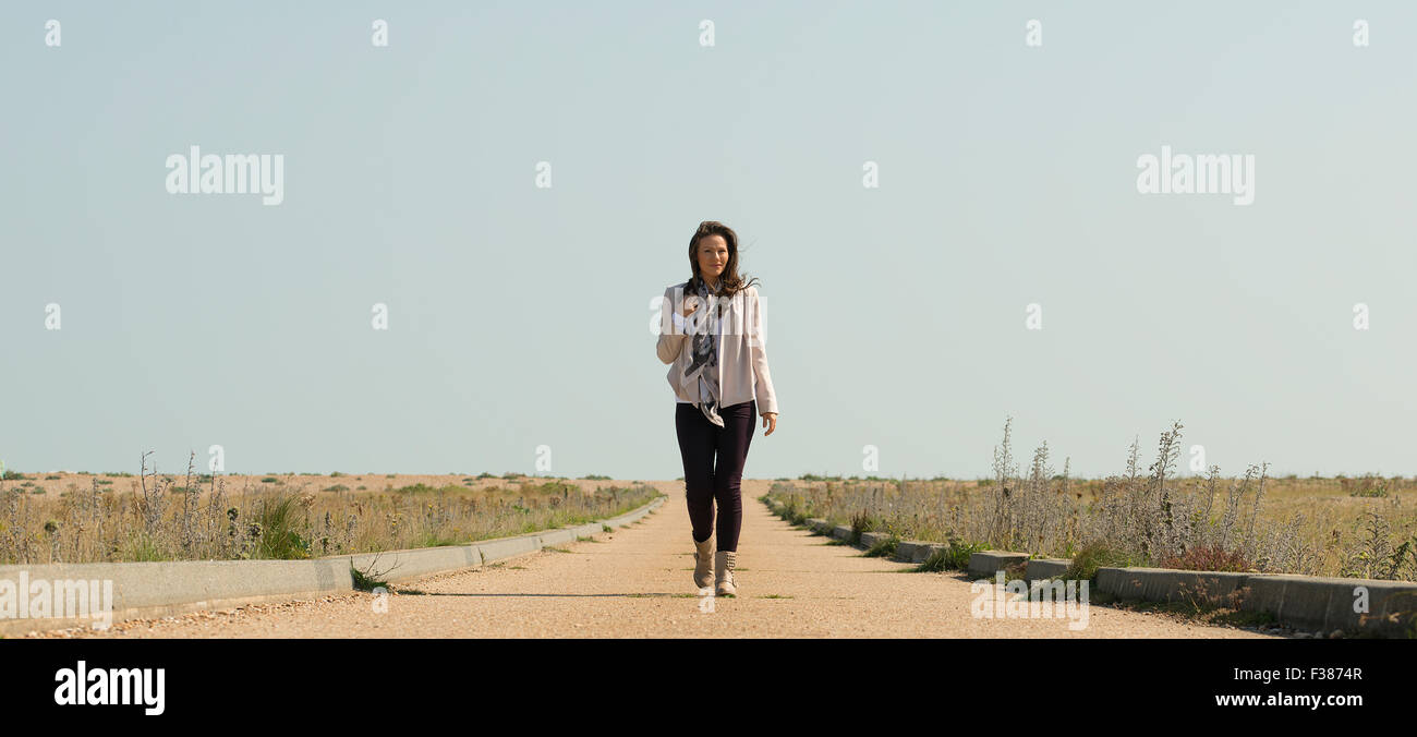 Young woman walking along a barren road, she is casually dressed and the sky is clear and blue. Stock Photo