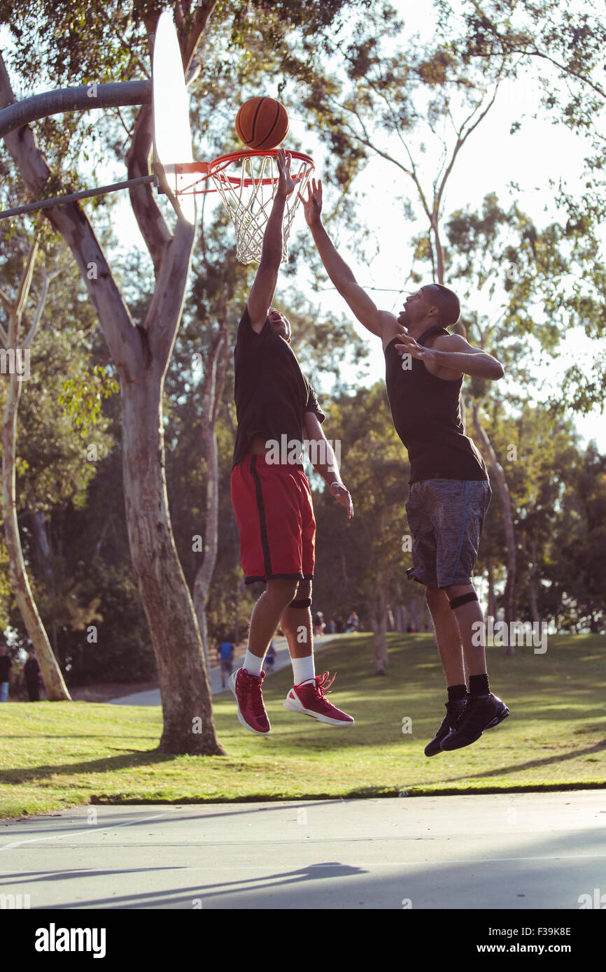 Two young men shooting basketball hoops in the park at sunset Stock