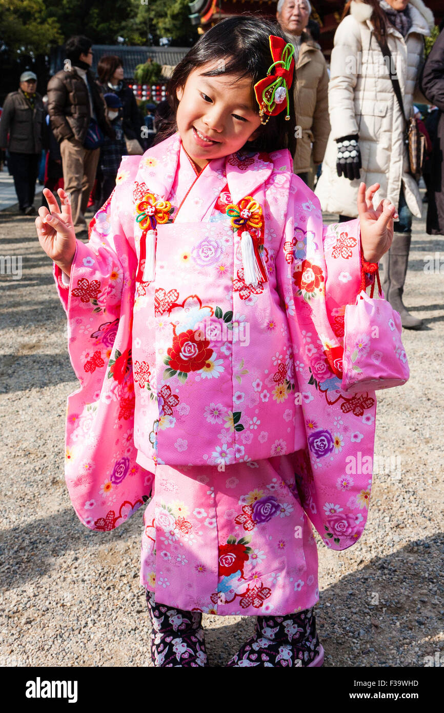 Japanese child, girl,4-5 years old, outdoors in winter sunlight, posing for viewer while wearing pink kimono. Smiling, hands in peace gesture. Stock Photo