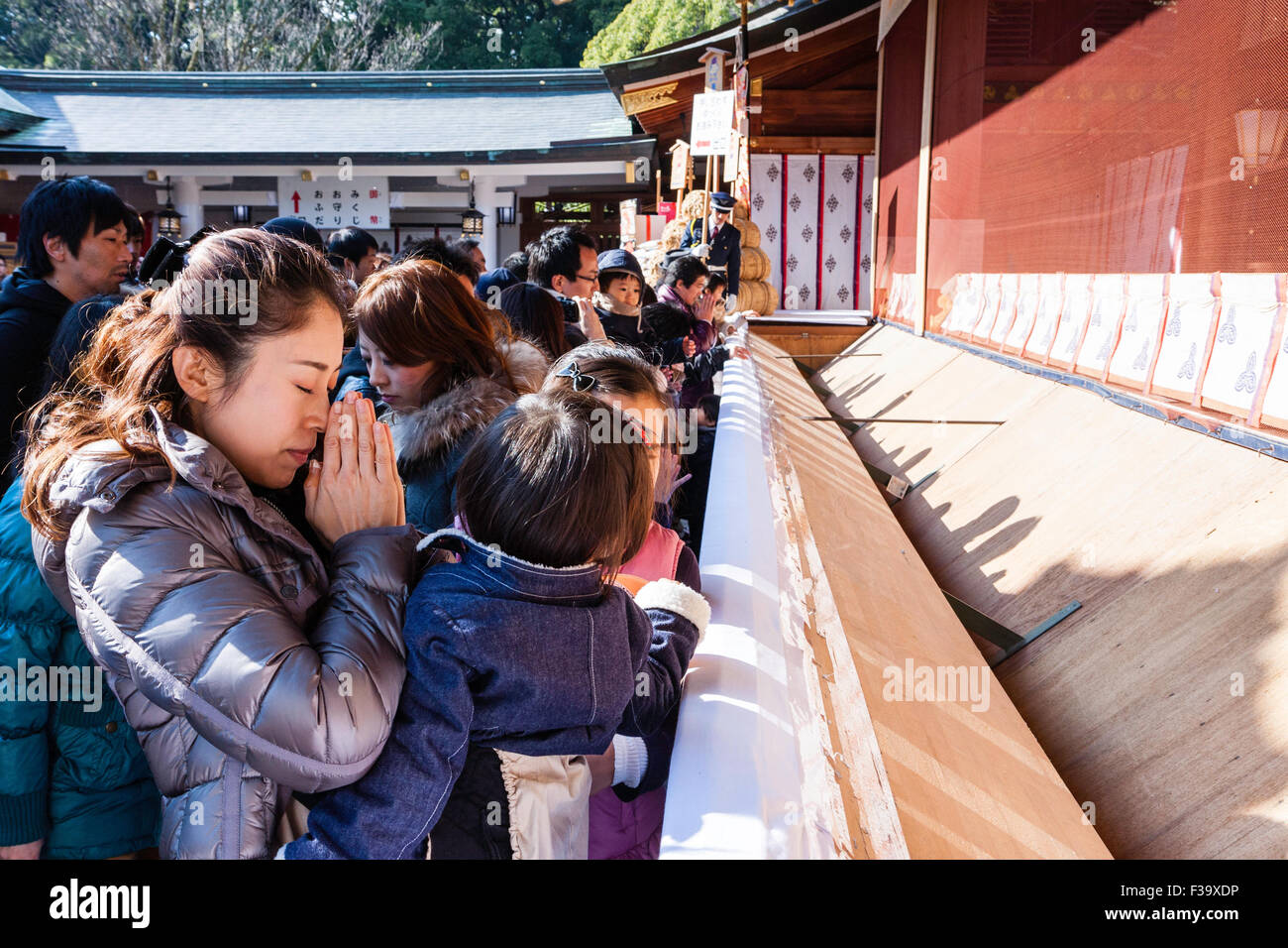 Japan, new year, winter. Japanese woman, mother, holding hands together and praying with two infant children at crowded Shinto shrine. Daytime. Stock Photo
