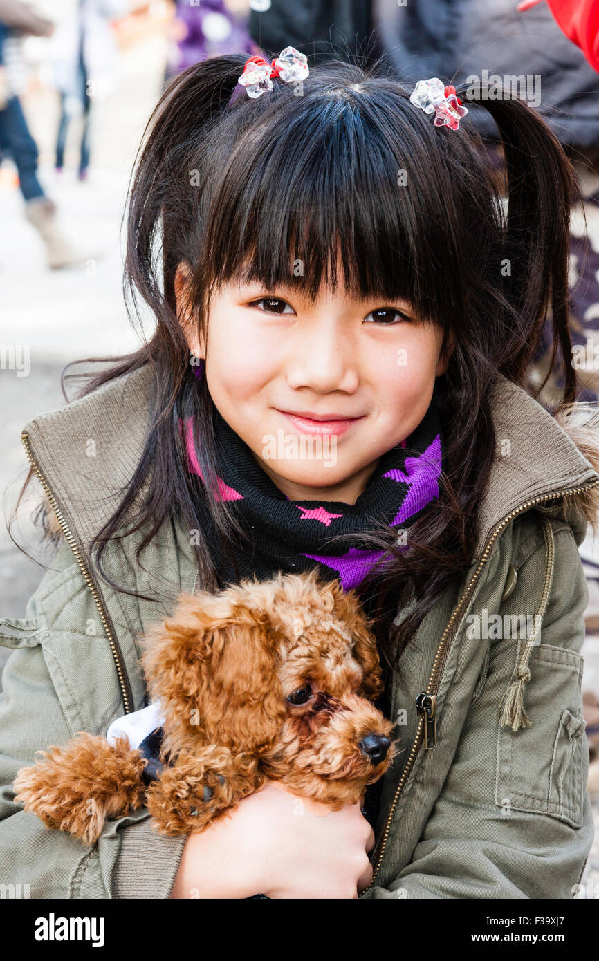 Japanese child, girl, 9-10 year old, in coat, smiling and posing for viewer while she cuddles and holds in both arms her pet dog. Eye-contact. Stock Photo