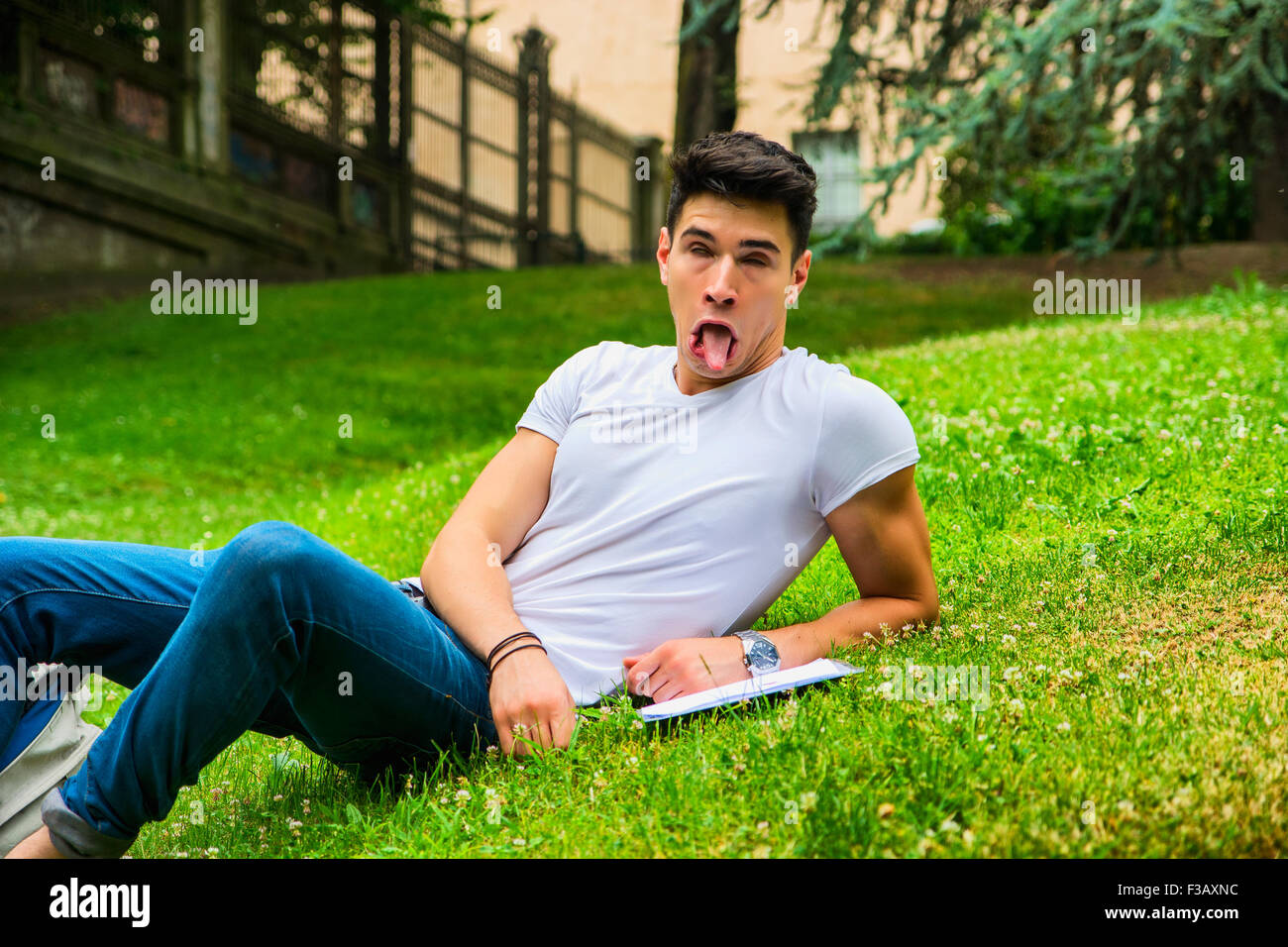 Young Male Student Doing Silly Face While Studying his Lessons, Lying on Grass in City Park Stock Photo