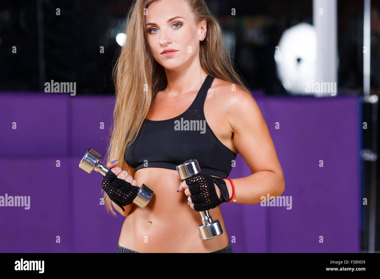 Strong young woman, with long hair, wearing in black top and breeches, holding dumbbells and looking at camera, on the sport equ Stock Photo