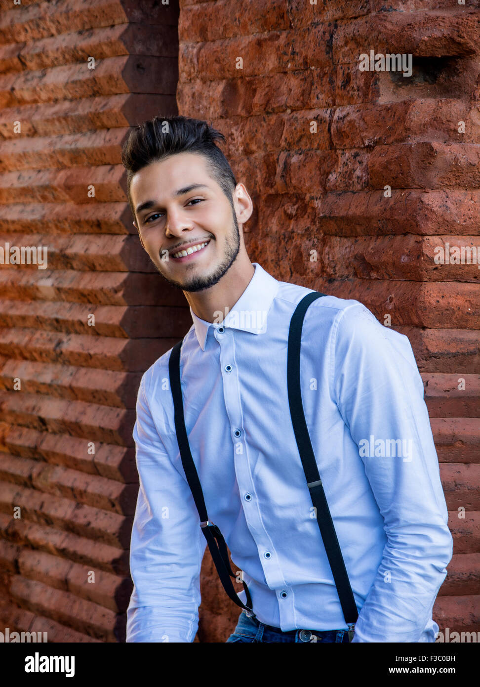 Attractive young man sitting against brick wall, looking at camera smiling. Three quarters shot Stock Photo