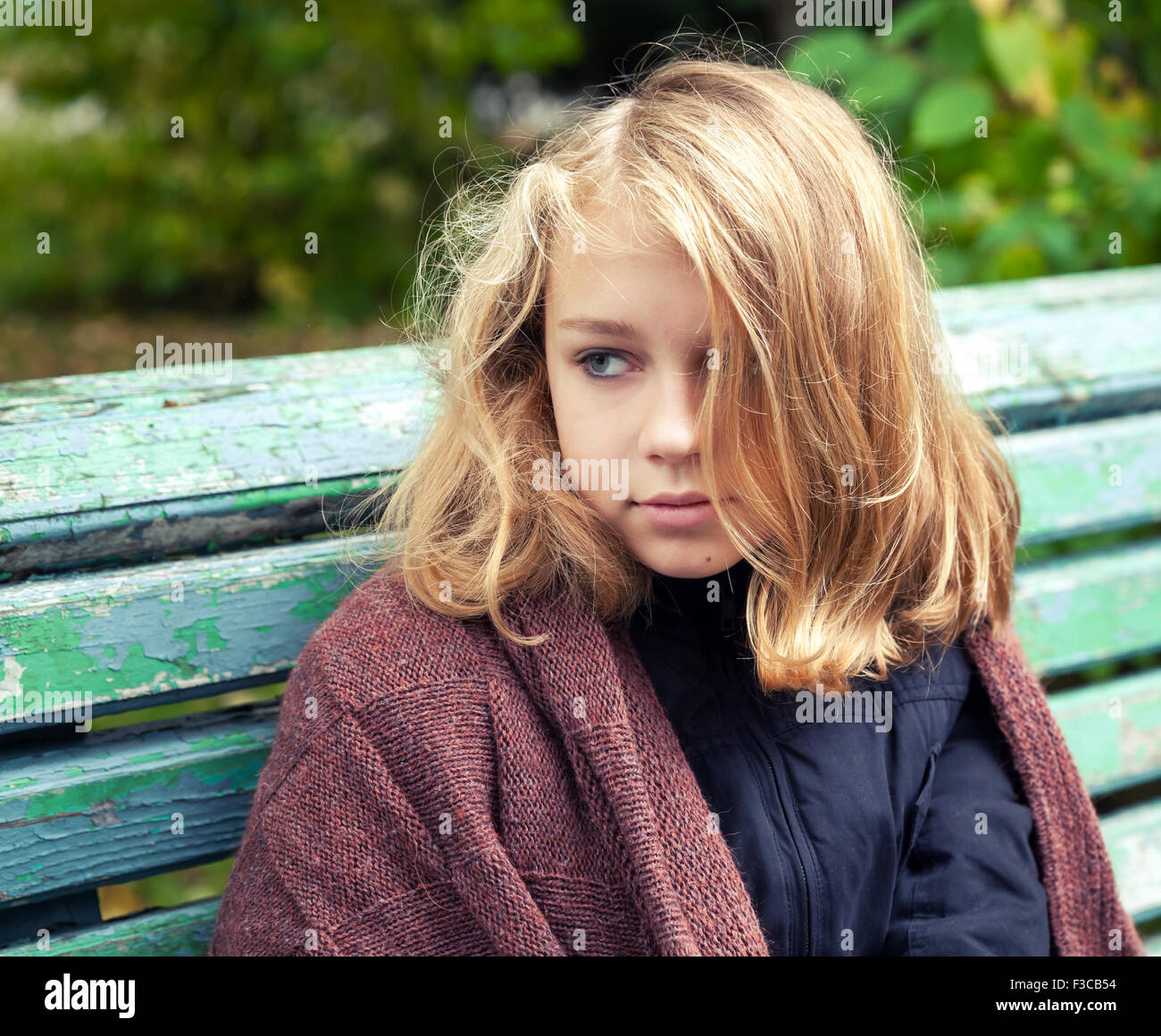 Beautiful Caucasian blond teenage girl in brown woolen plaid sitting on old green bench in autumn park Stock Photo