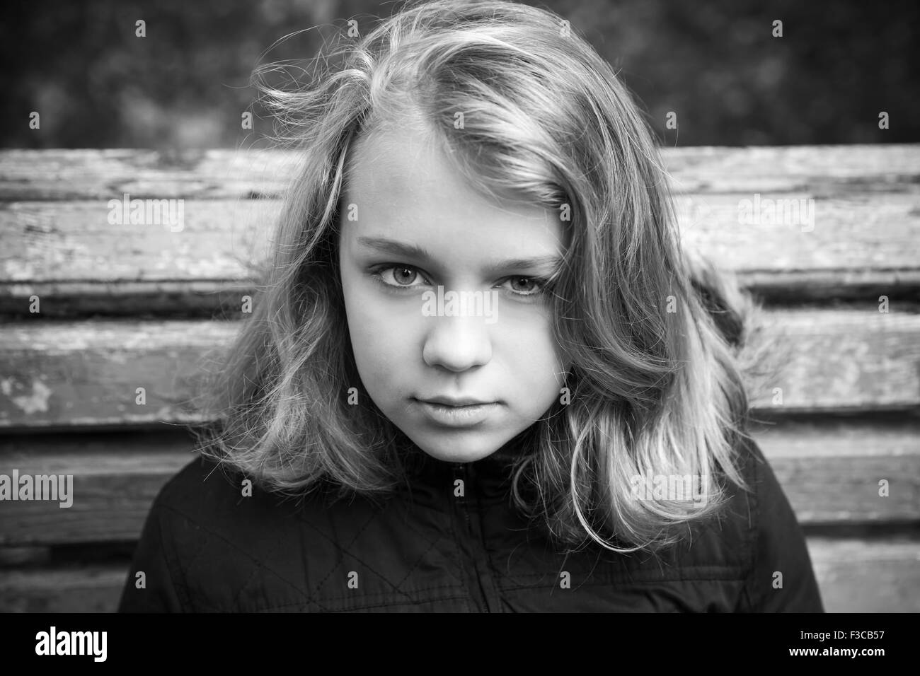 Beautiful Caucasian blond teenage girl in black sitting on old wooden bench in autumn park, monochrome photo Stock Photo