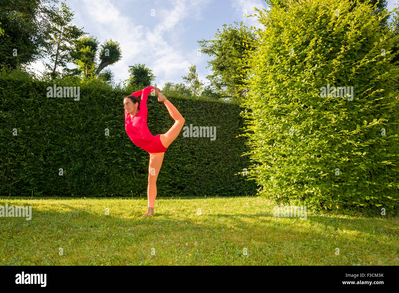 Young woman, wearing a red-orange body suit, is practising Hatha-Yoga outdoor between trees, showing the pose: natarajasana, Lor Stock Photo