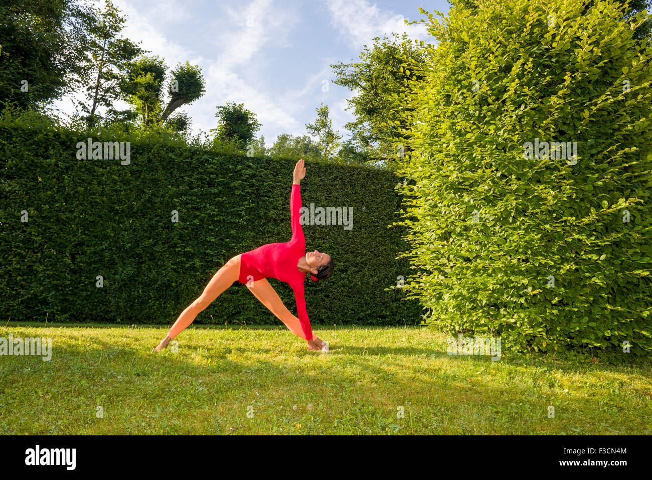 Young woman, wearing a red-orange body suit, is practising Hatha-Yoga outdoor between trees, showing the pose: trikonasana, tria Stock Photo