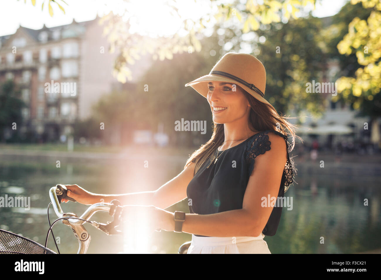 Portrait of happy young woman at the city park walking along a pond with her bicycle. European female model wearing hat looking Stock Photo