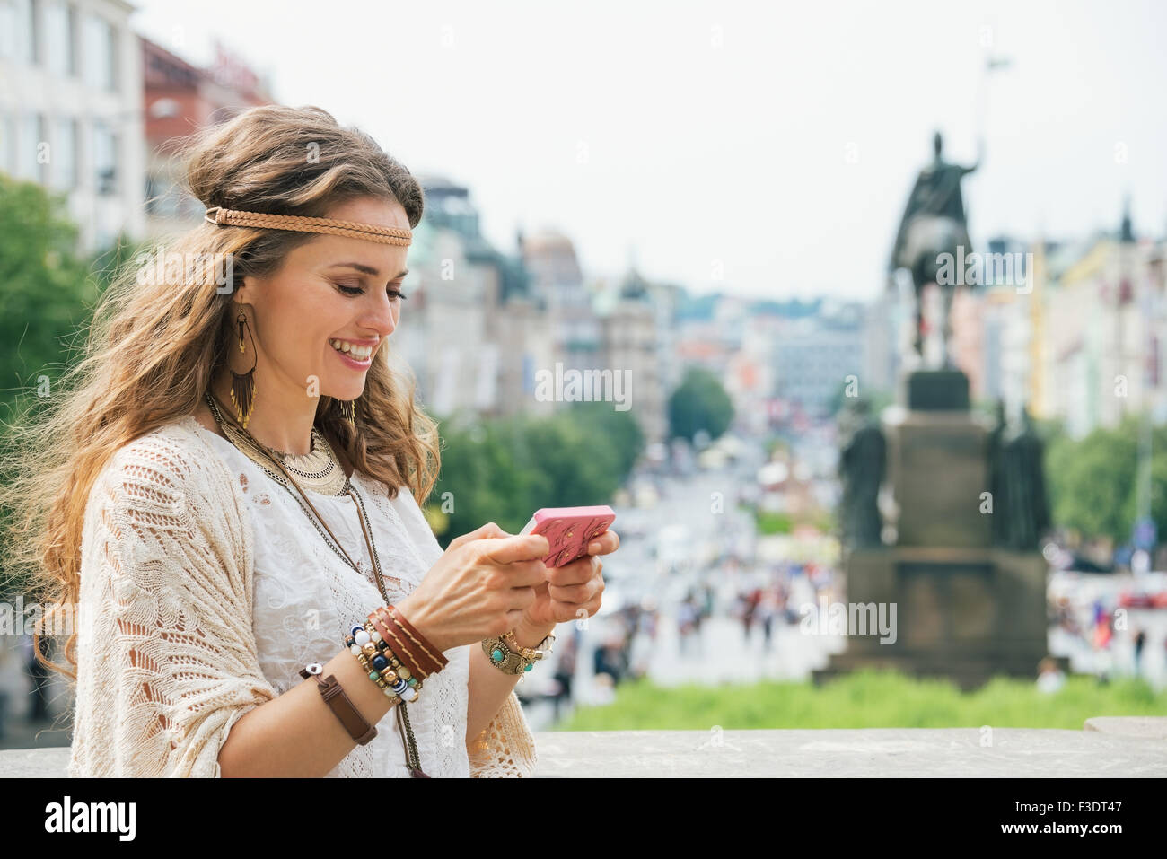 Smiling young longhaired bohemian young lady writing sms on mobile on Wenceslas Square in Prague. In the background Saint Wences Stock Photo