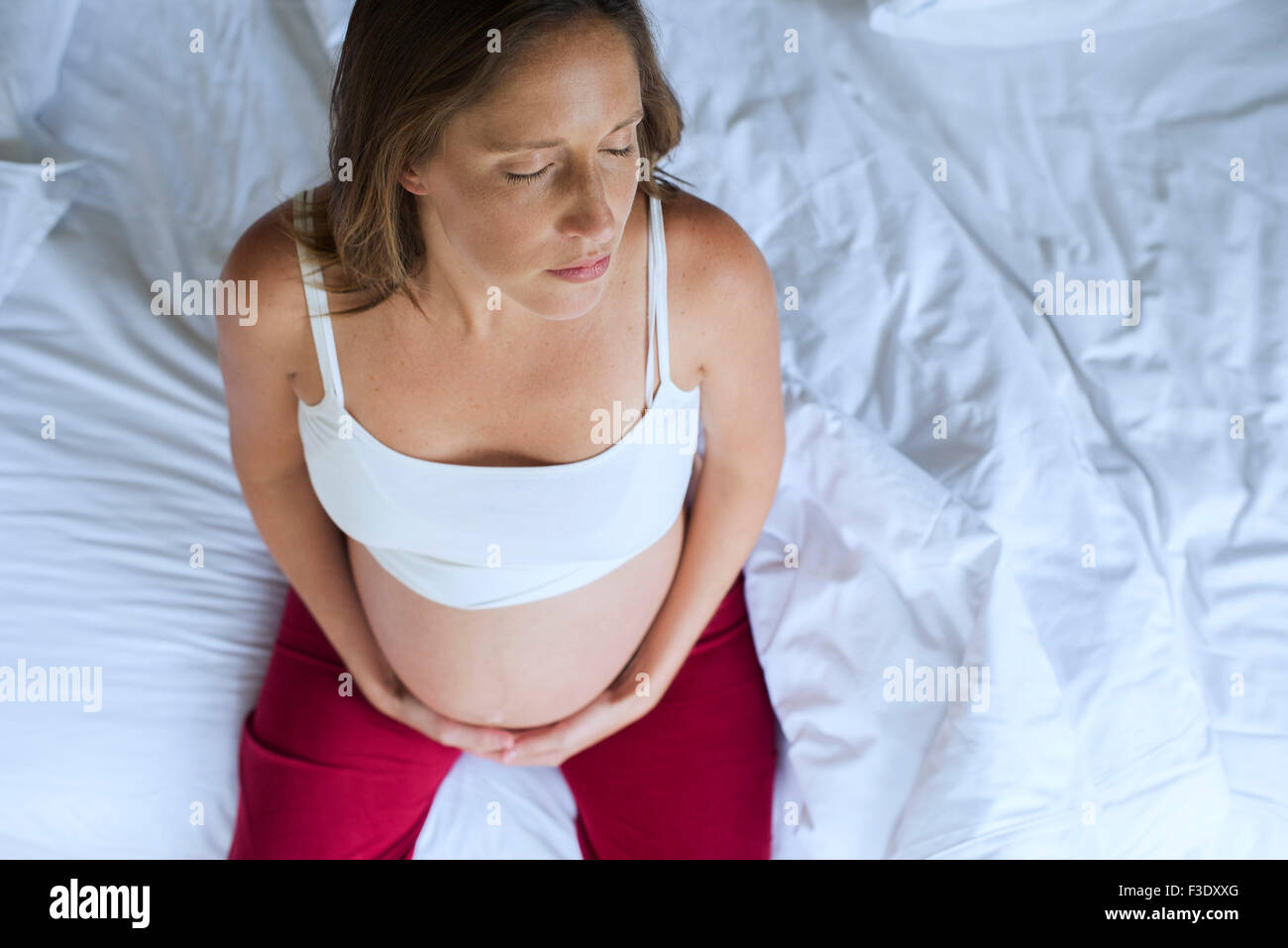 Pregnant woman sitting on bed with hands on stomach and eyes closed Stock Photo
