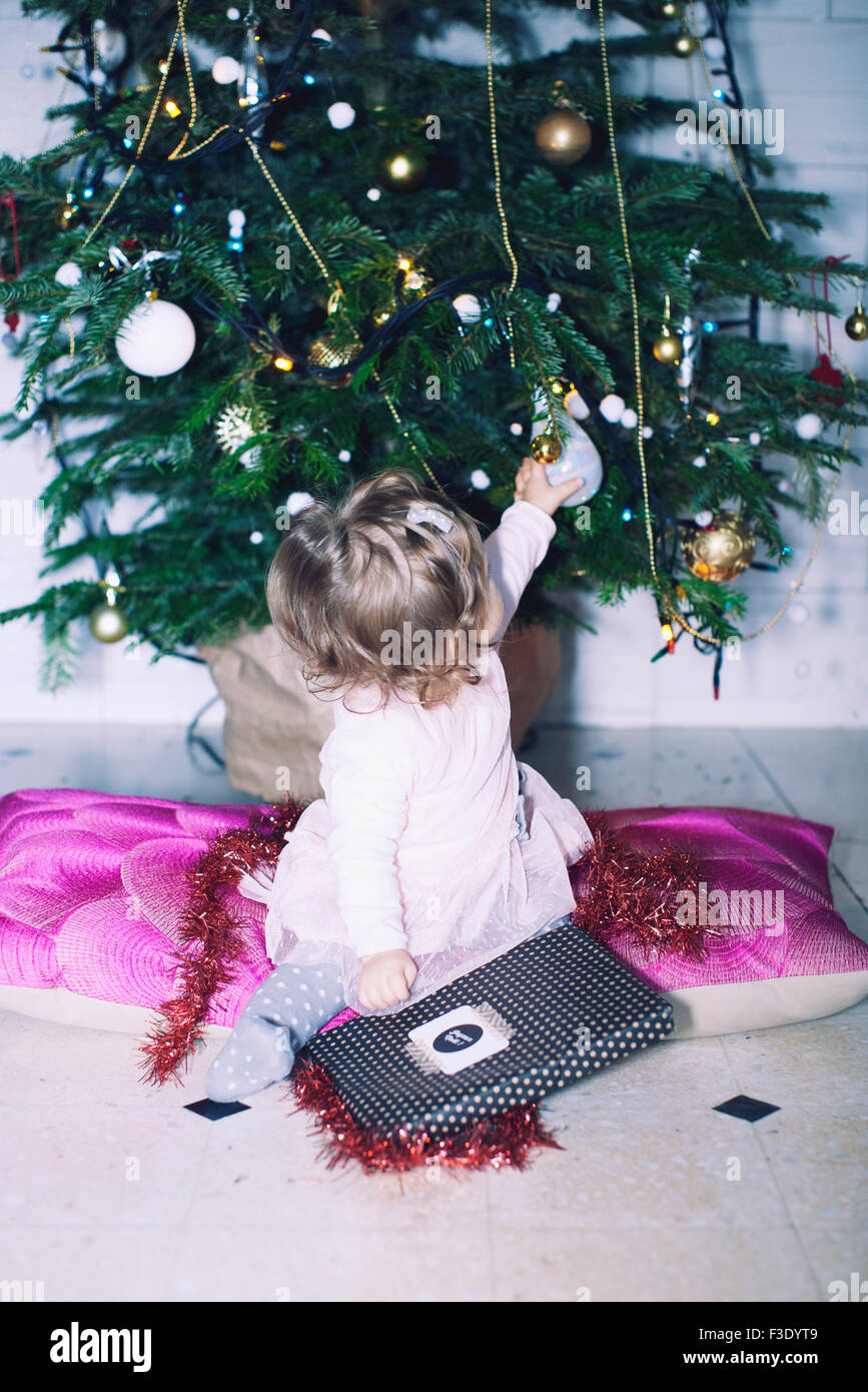 Baby girl sitting in front of Christmas tree with gift on lap, looking over shoulder to touch ornament Stock Photo