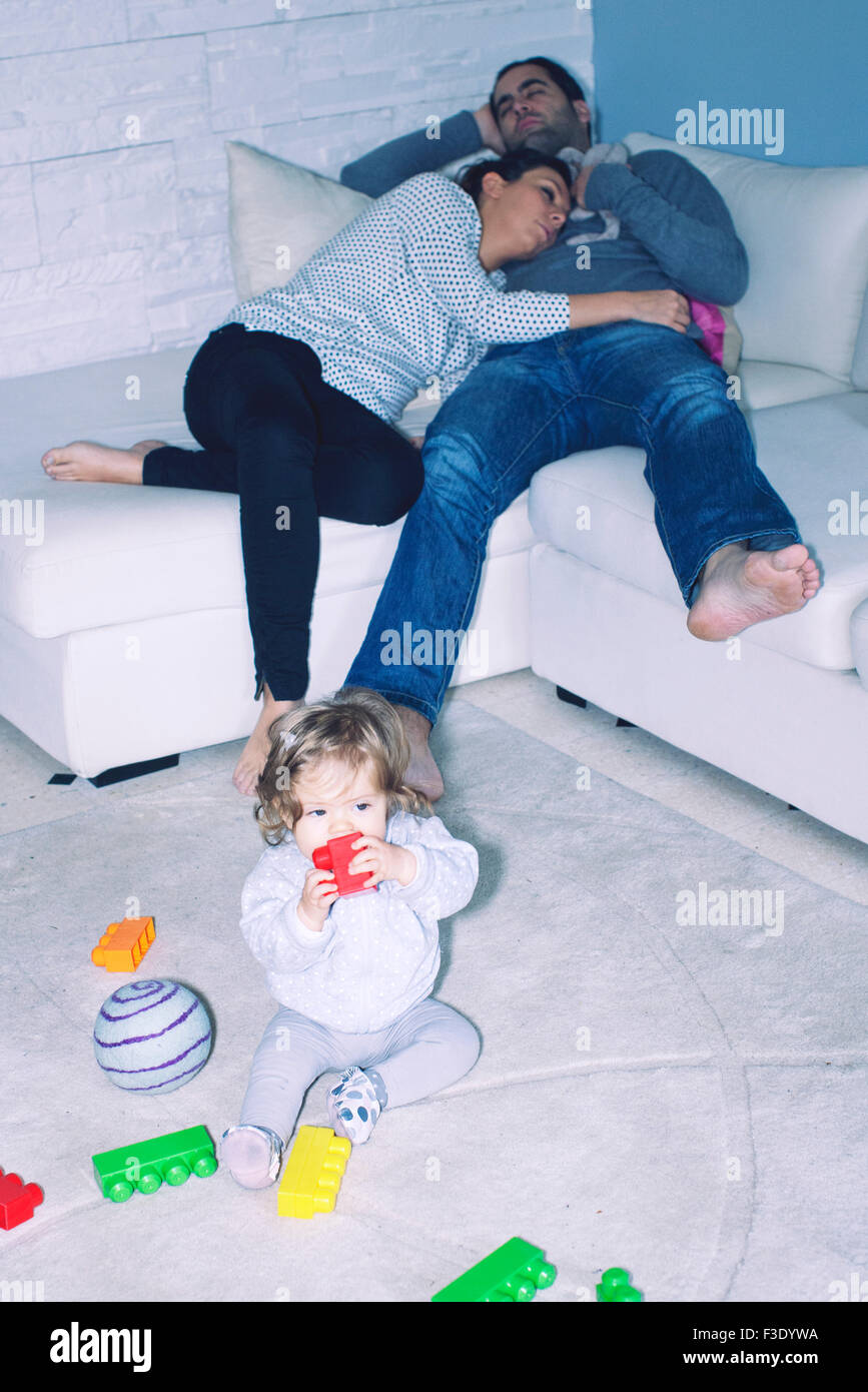 Parents sleeping on sofa while baby girl plays with toys on the floor Stock Photo