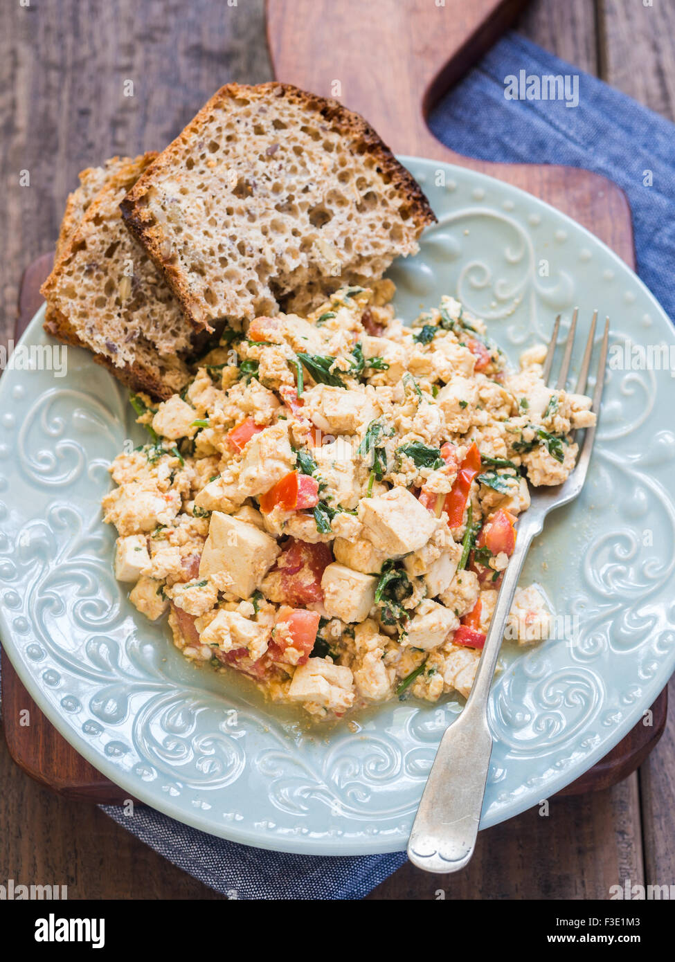 Vegan tofu scramble with tomato and green herbs served with wholegrain homemade bread. Wooden background. Stock Photo