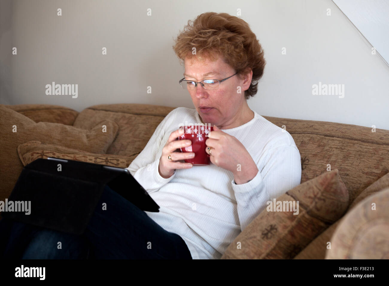 A middle aged woman relaxing at home with a cup of coffee whilst browsing the internet using a tablet ipad computer Stock Photo