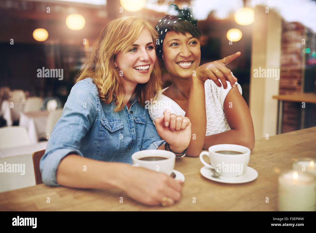 Happy girl friends enjoying coffee together in a coffee house sitting pointing and smiling with amusement as they watch somethin Stock Photo