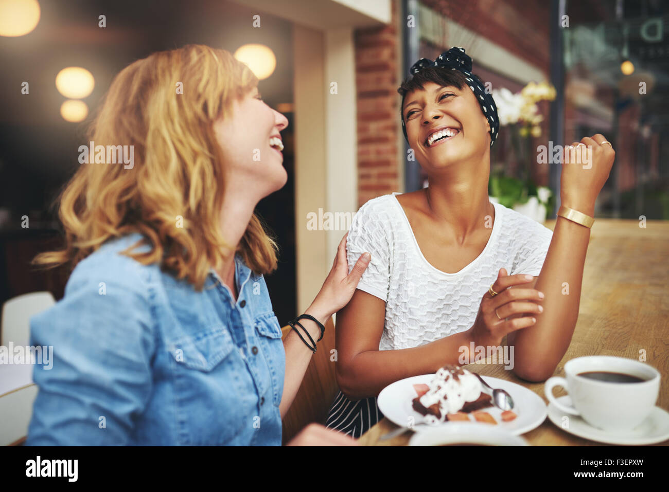 Two multiethnic young female friends enjoying coffee together in a restaurant laughing and joking while touching to display affe Stock Photo
