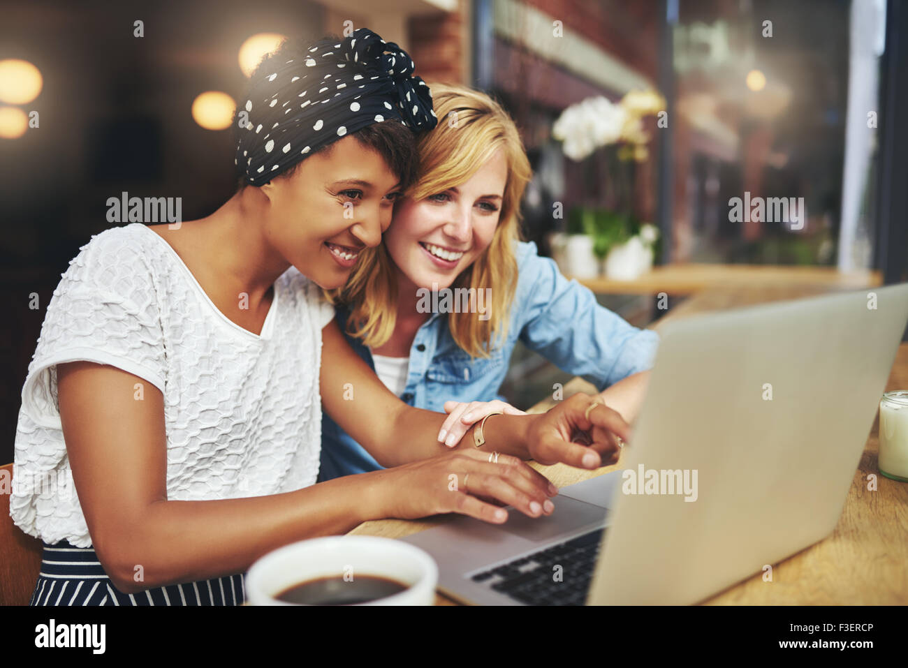Two multiracial young female friends surfing the internet together on a laptop as they sit in a cafeteria enjoying a cup of coff Stock Photo