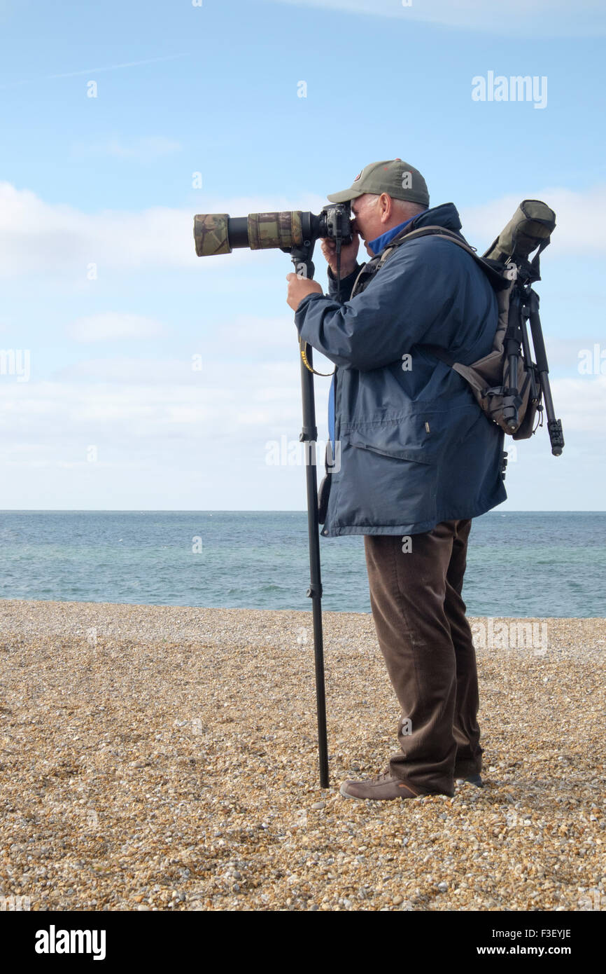 A man birdwatching on a pebble beach at Salthouse, Norfolk, with a large camera and a monopod. Stock Photo