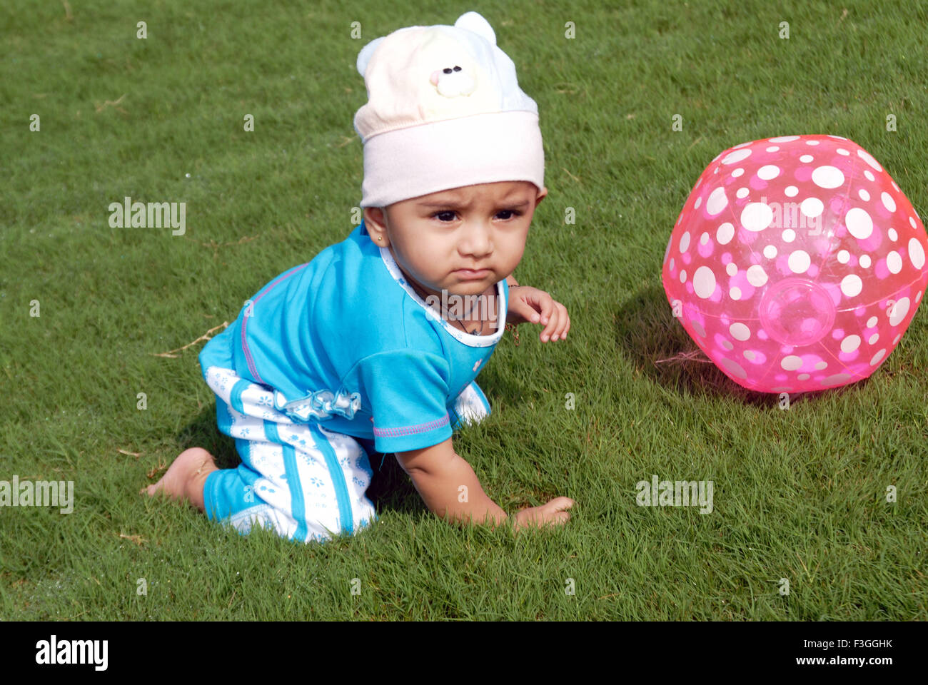 Baby crawling wearing white cap in blue dress playing with pink plastic ball on green grass MRr#152 Stock Photo