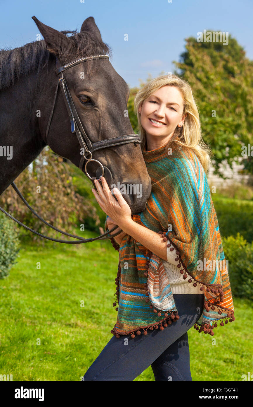Beautiful happy woman wearing poncho, smiling and leading her horse in sunshine Stock Photo
