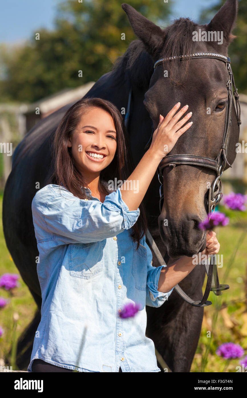 Beautiful happy Asian Eurasian young woman or girl wearing denim shirt, smiling and leading her horse in sunshine Stock Photo