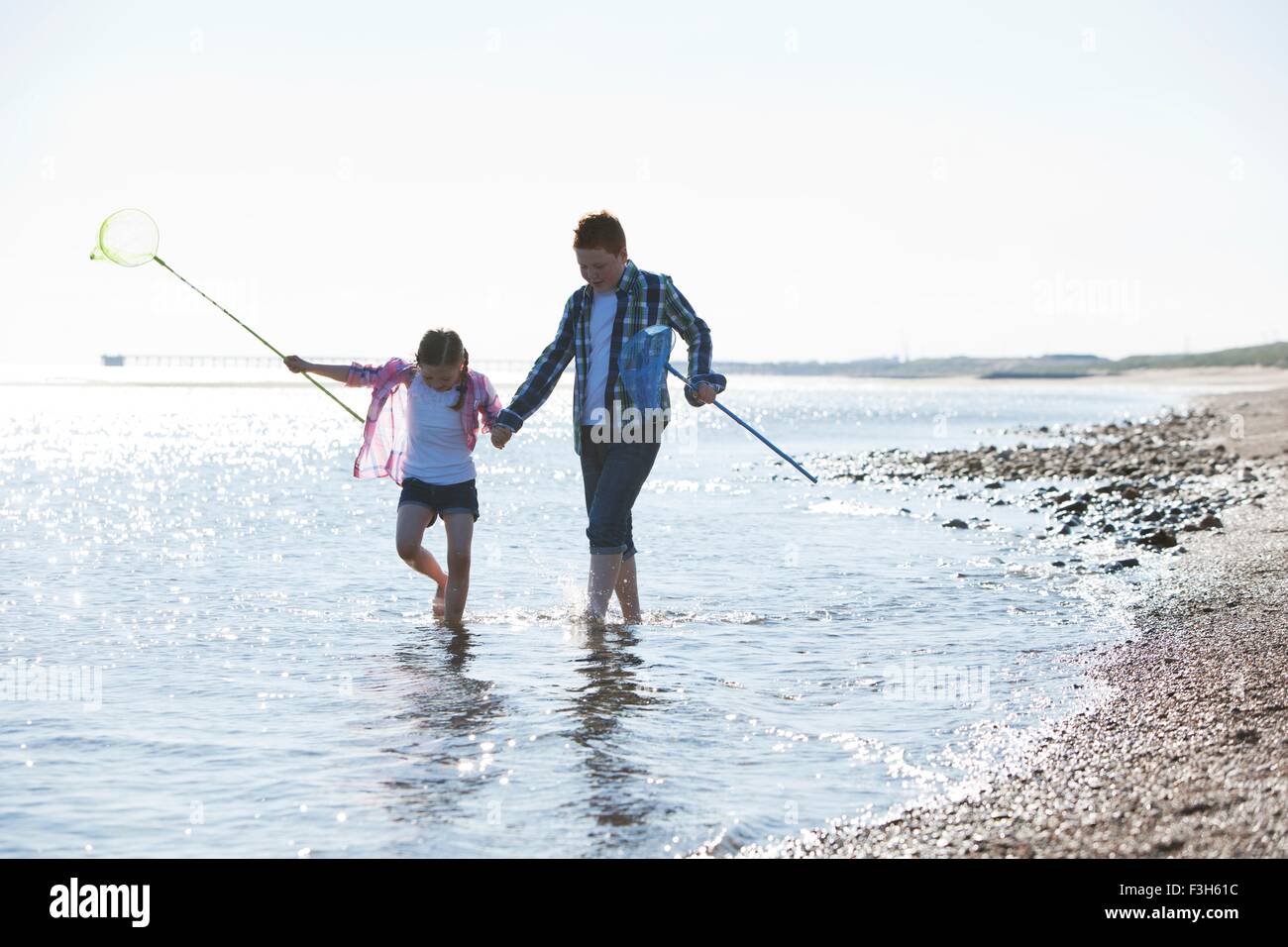 Girl and boy paddling in water holding hands, carrying fishing nets, looking down Stock Photo