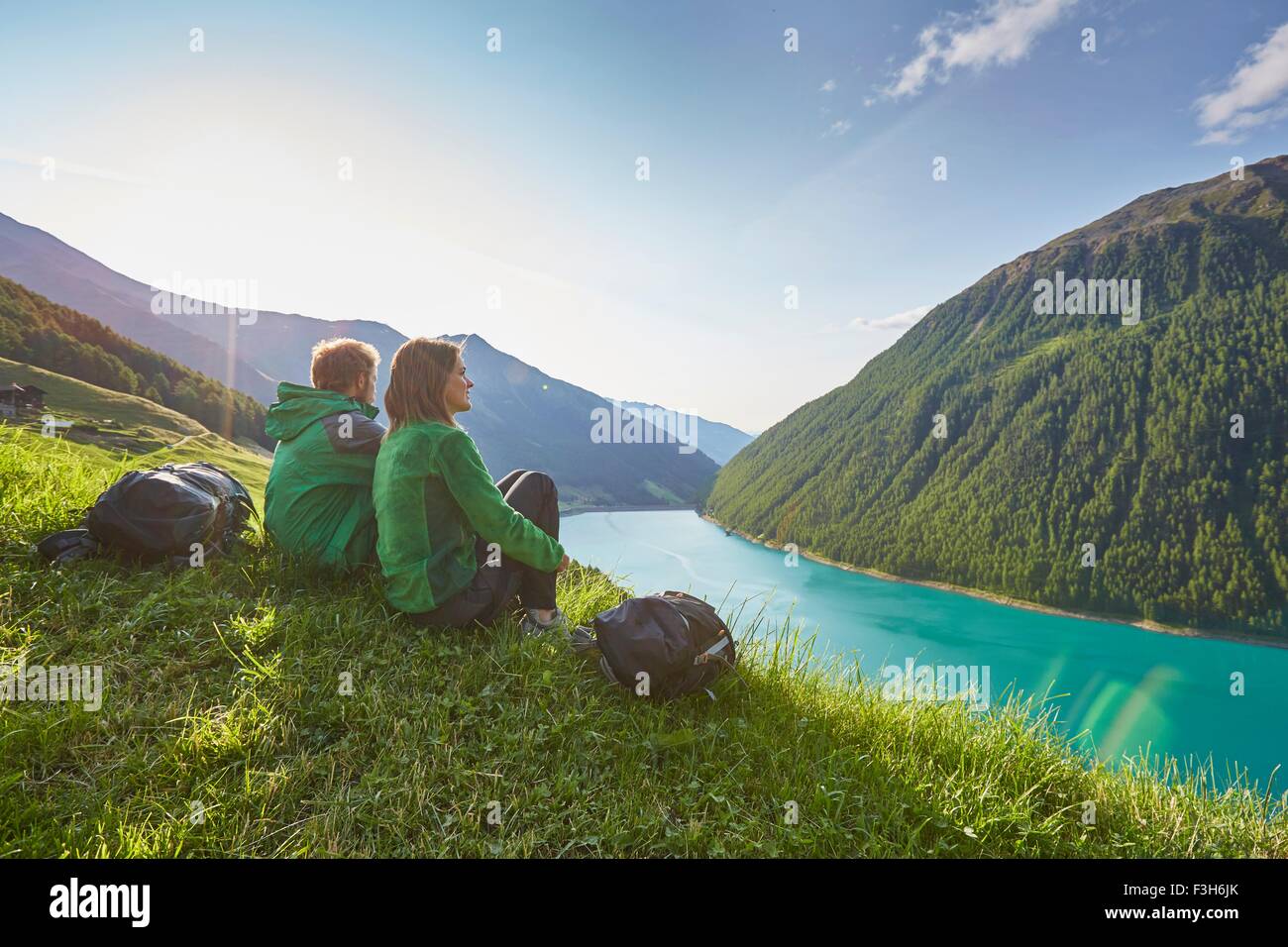 Young couple sitting  looking out over Vernagt reservoir and Finailhof farmhouse, Val Senales, South Tyrol, Italy Stock Photo