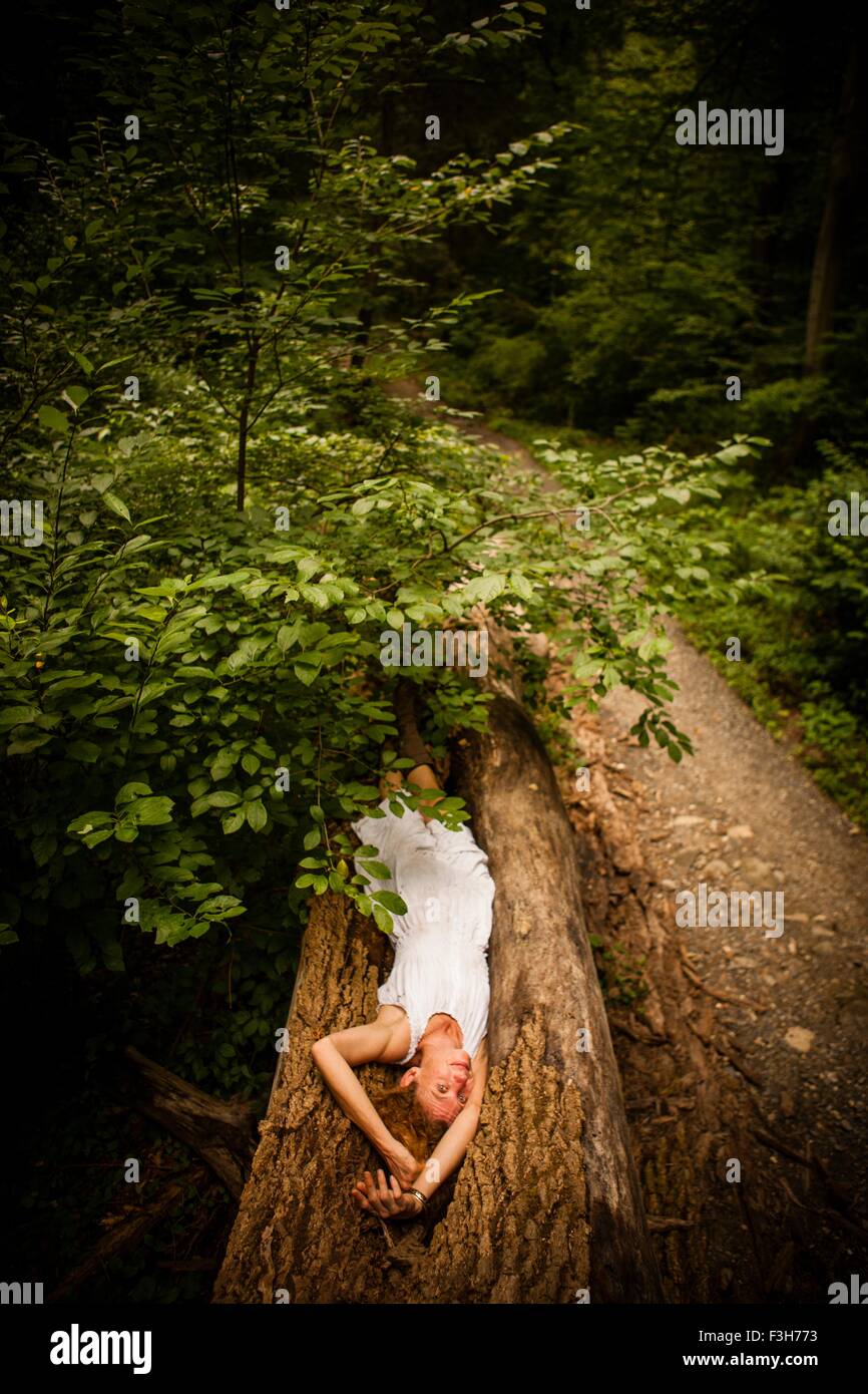 Mid adult woman wearing white dress lying on fallen tree arms raised above head Stock Photo