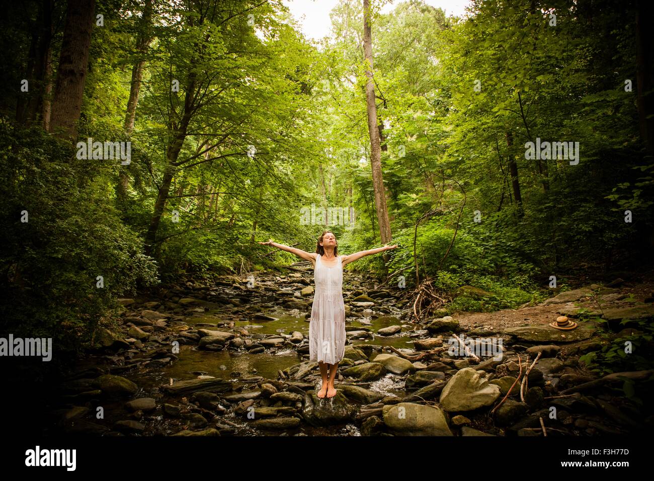 Mid adult woman standing barefoot on rocky riverbed wearing white dress, arms raised open looking up Stock Photo