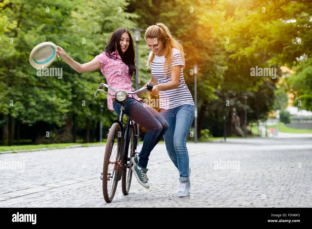 Young woman holding hat riding side saddle on bicycle with friend pushing Stock Photo