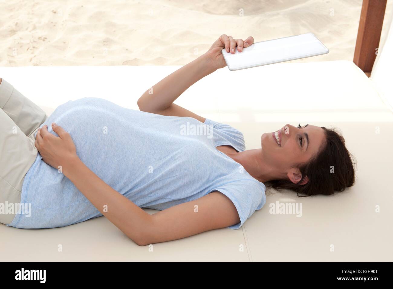Pregnant mid adult woman lying on beach daybed looking up at digital tablet Stock Photo