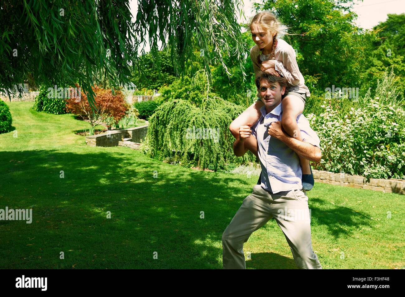 Mature man running with daughter on his shoulders in garden Stock Photo