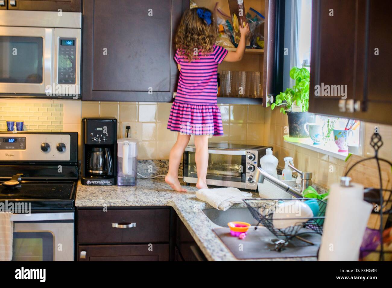 Young girl standing on kitchen work surface, looking for treats in cupboard, rear view Stock Photo