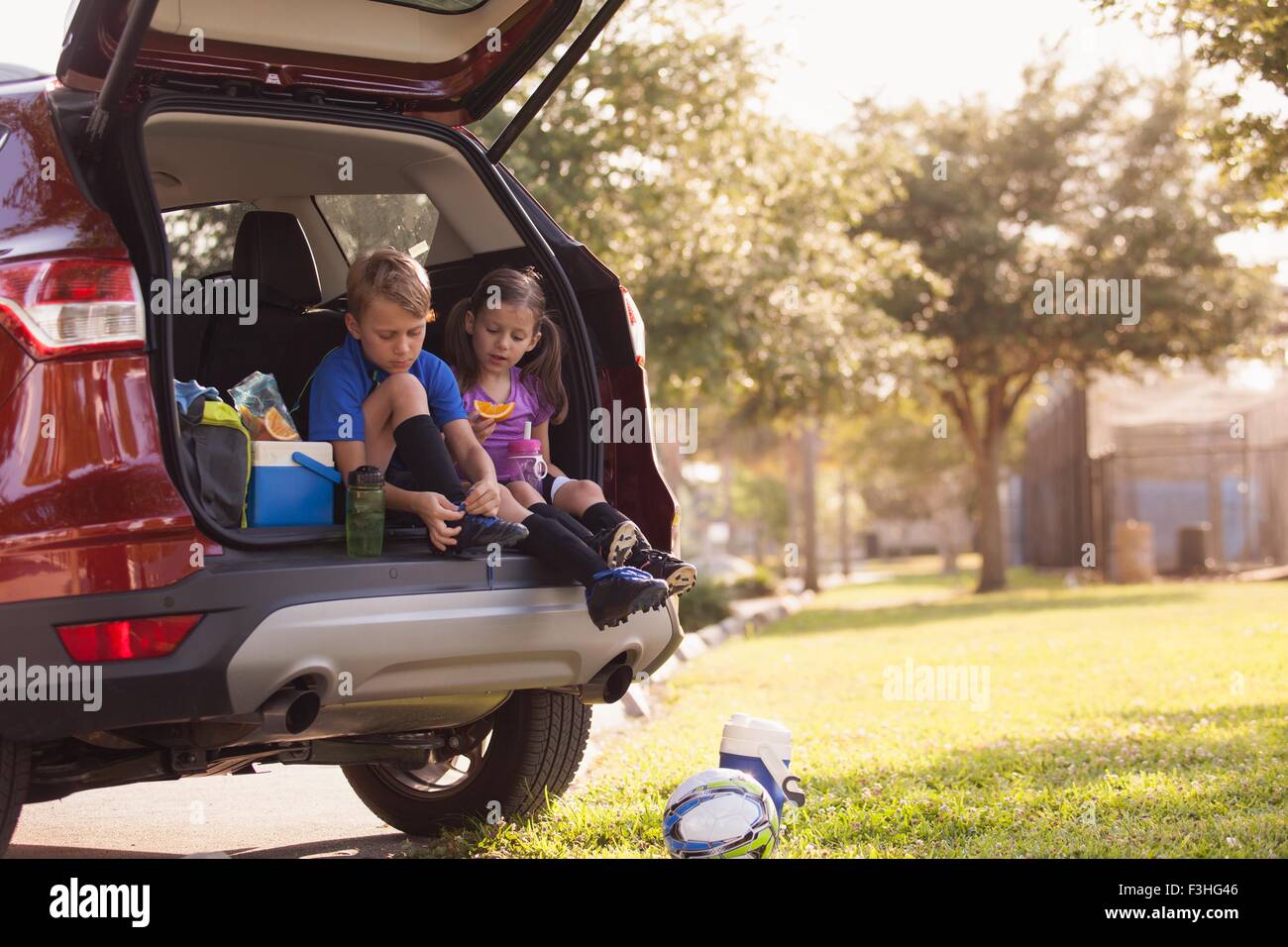 Boy and younger sister preparing for football practice whist sitting in car boot Stock Photo